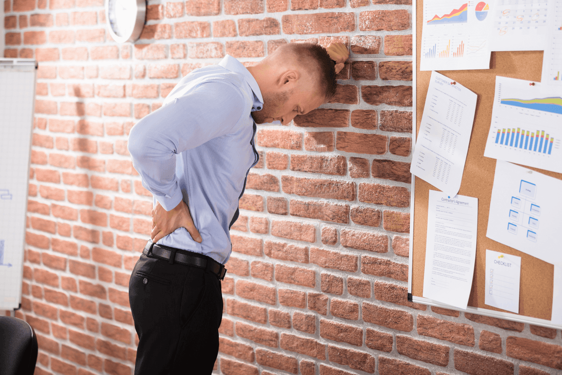 A man facing and leaning on the brick holding his lower back at his office who appears to be suffering from excruciating sciatic pain.